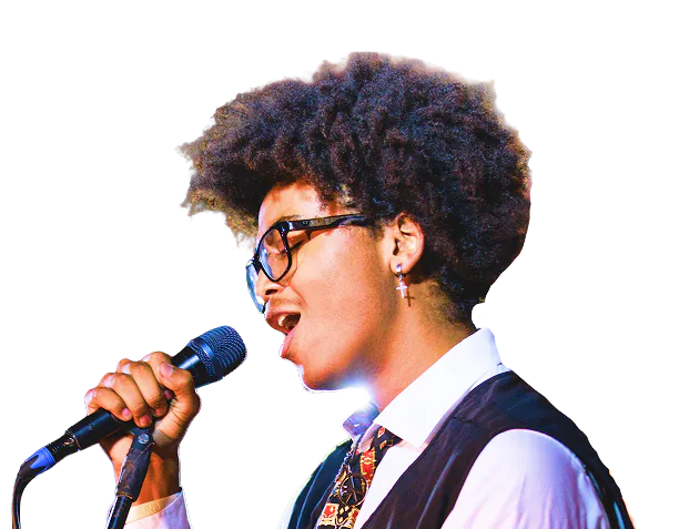 Young person with curly hair, glasses, and a cross earring singing into a microphone, wearing a white shirt, patterned tie, and dark vest against a black background.